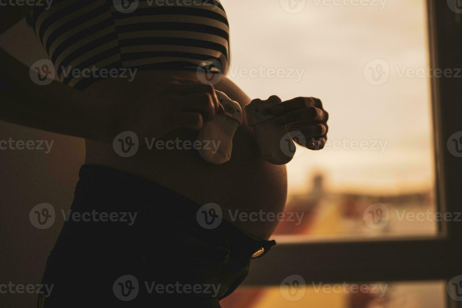 embarazada mujer en pie junto a ventana y participación bebé medias. silueta de embarazada mujer barriga. foto
