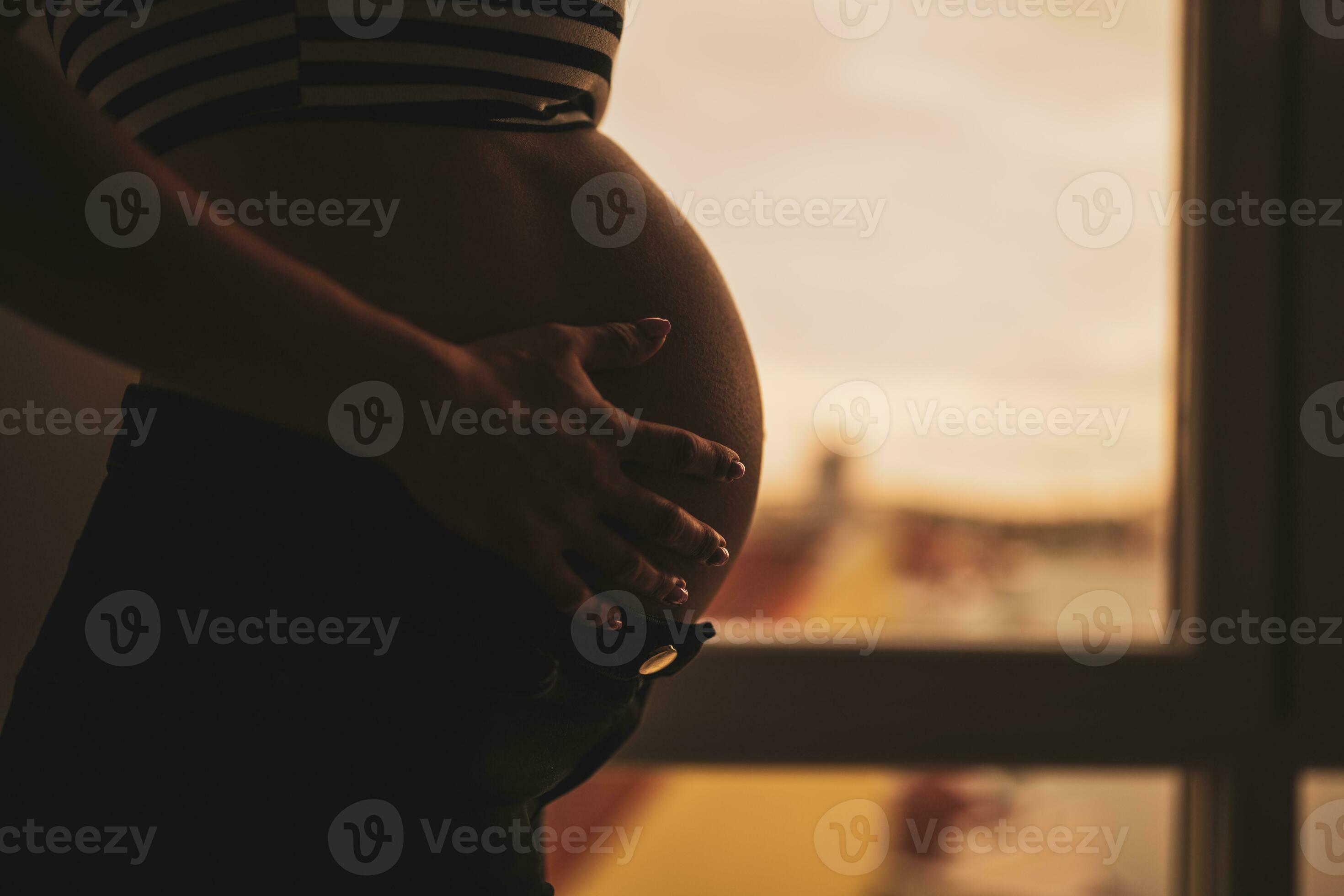 Close up of pregnant woman standing beside window and holding her belly