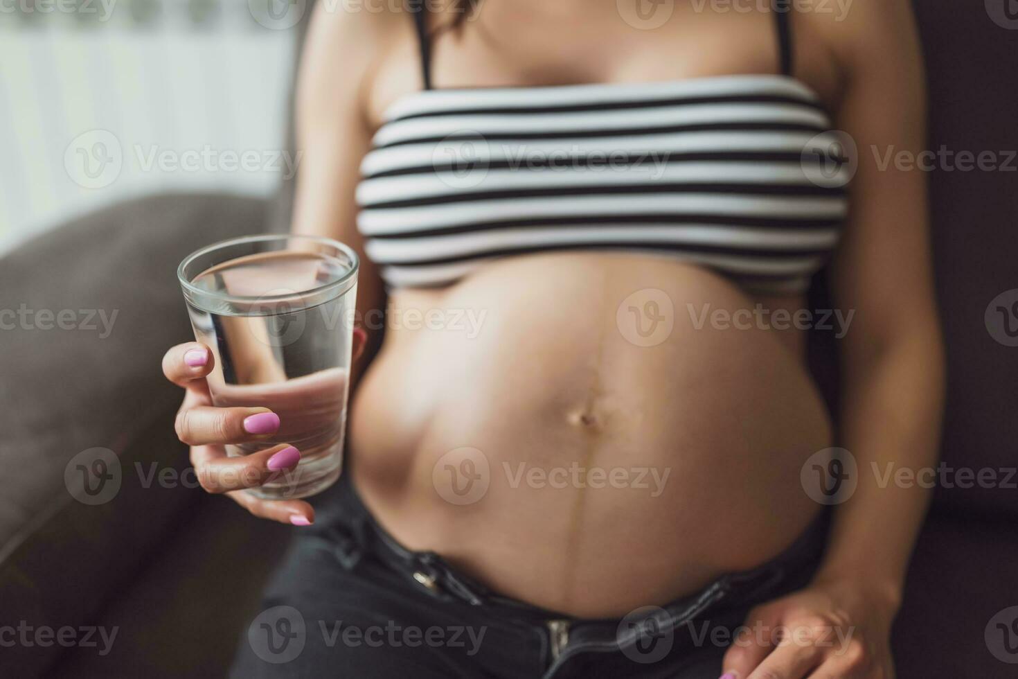 embarazada mujer relajante a hogar. ella es sentado en cama y Bebiendo agua. foto