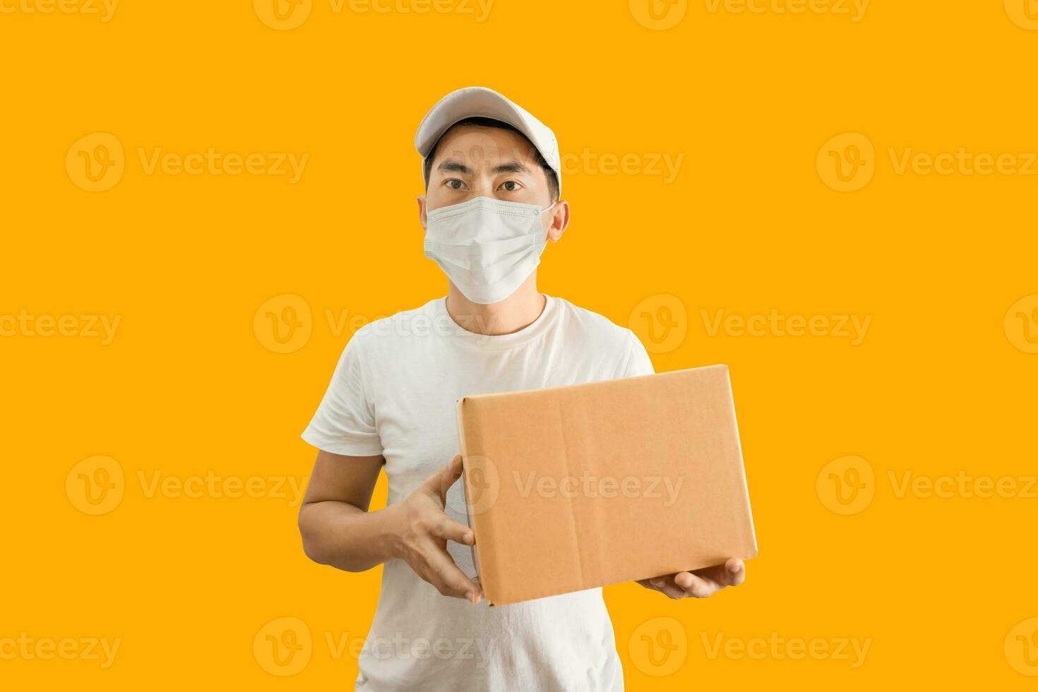 Young Asian delivery man wearing cap and white blank t-shirt holding parcel post box isolated on yellow background. express delivery service concept. photo