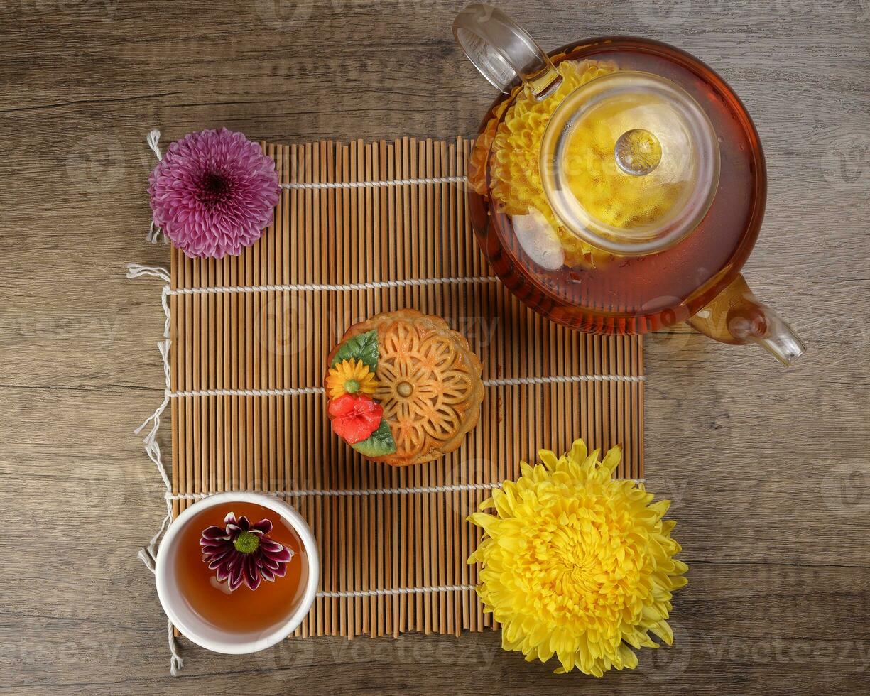 Colorful flower decorated moon cake Chinese mid autumn festival tea in small teacup glass teapot daisy chrysanthemum mum flower red yellow pink purple violet bamboo food mat on yellow background photo