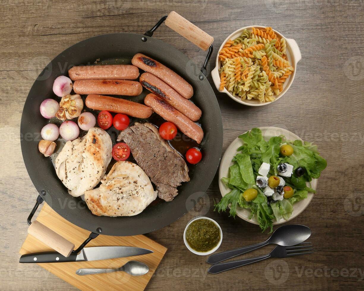 Mix meet chicken beef steak sausage tomato onion garlic fry grill oil metal fry pan colourful pasta green salad with cheese olive mint basil pesto sauce board knife fork spoon rustic wooden table photo