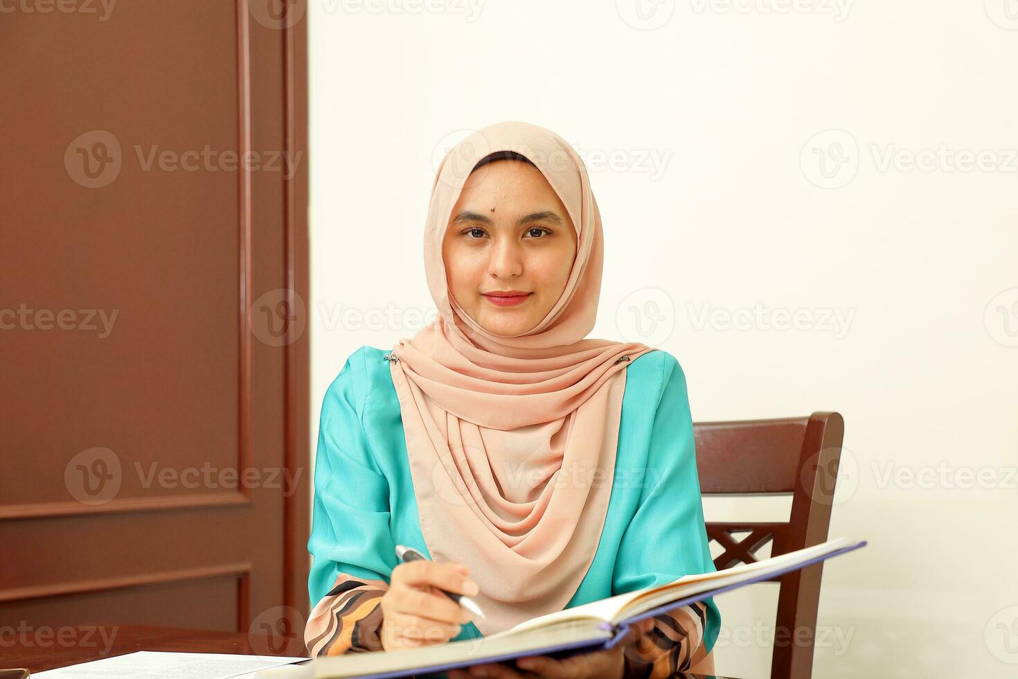young Asian Malay Muslim woman wearing headscarf at home office student sitting at table read write book document note study smile happy look at camera photo