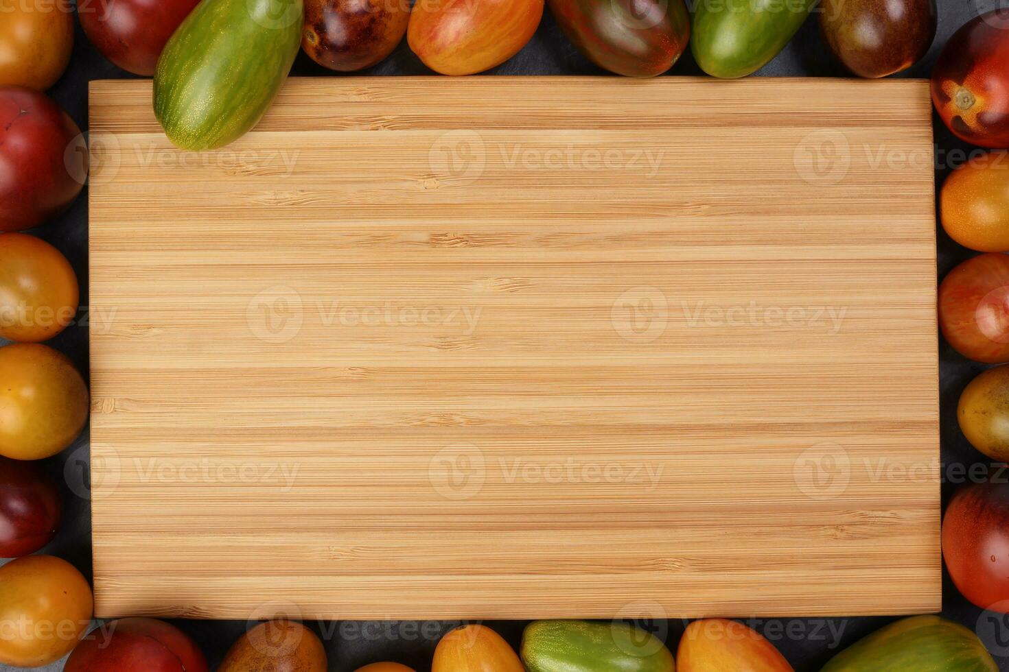 Red orange yellow green tomato mix variety on around wooden chopping board frame copy text space over black slate stone background photo