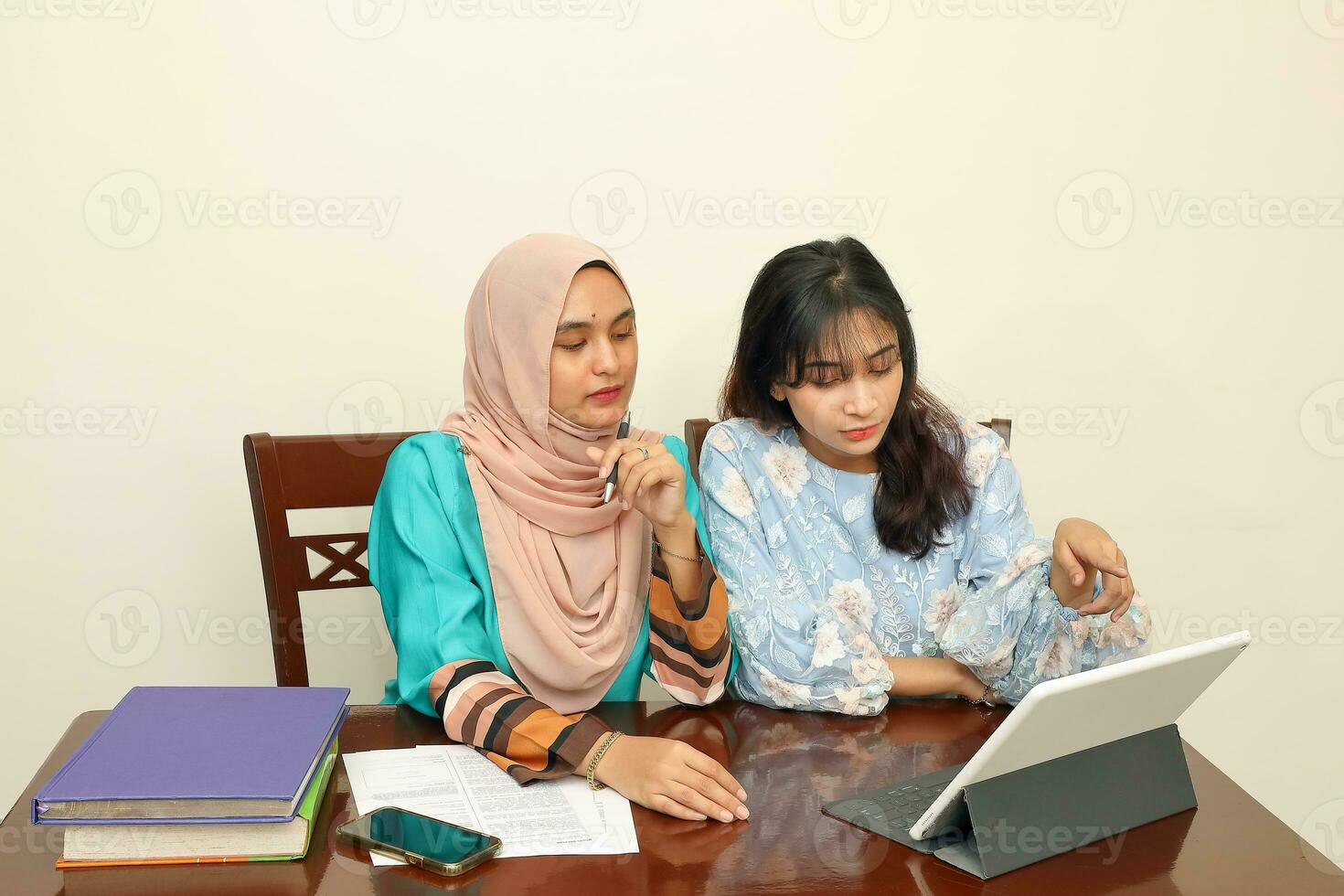Two young Asian Malay Muslim woman wearing headscarf at home office student sitting at table talk mingle look at computer book document study discuss read type write smile happy photo