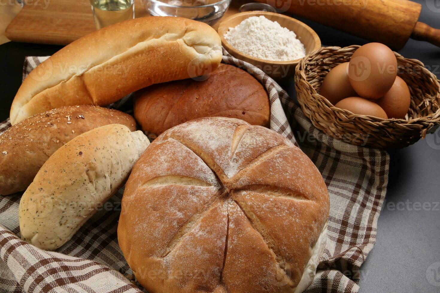 Freshly baked bread loaf bun roll round long mix verity wrapped in checkered kitchen fabric napkin towel wheat flower oil water salt eggs rolling pin slate stone over black background photo