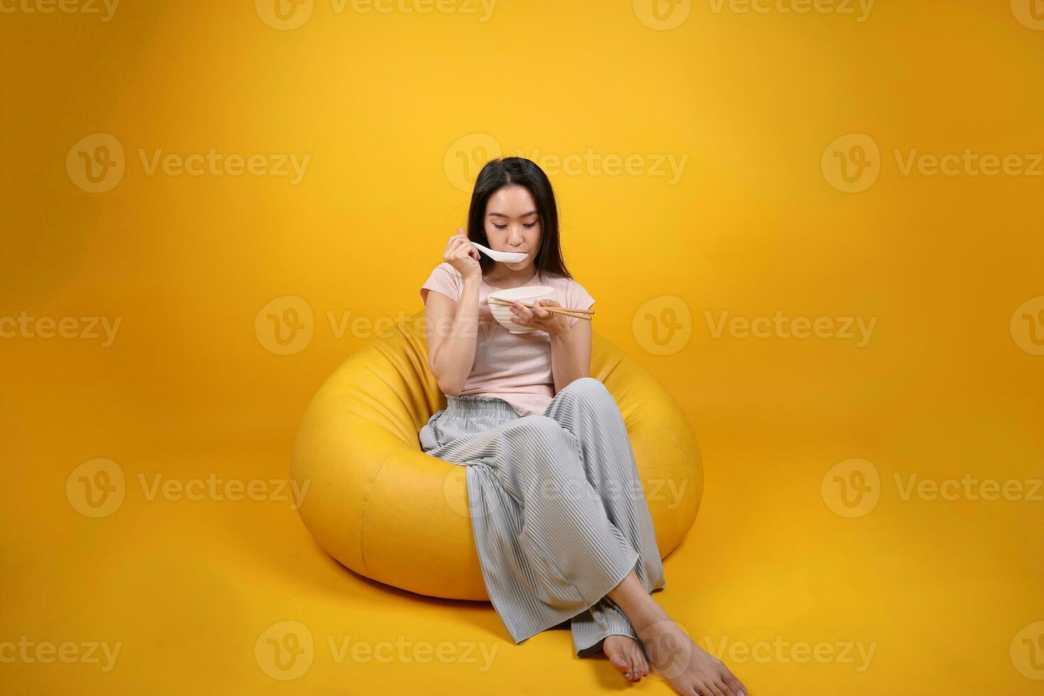 Beautiful young south east Asian woman holding empty chopstick soup spoon bowl pretend acting posing see eat taste smell feed offer satisfaction yummy siting on beanbag seat yellow orange background photo