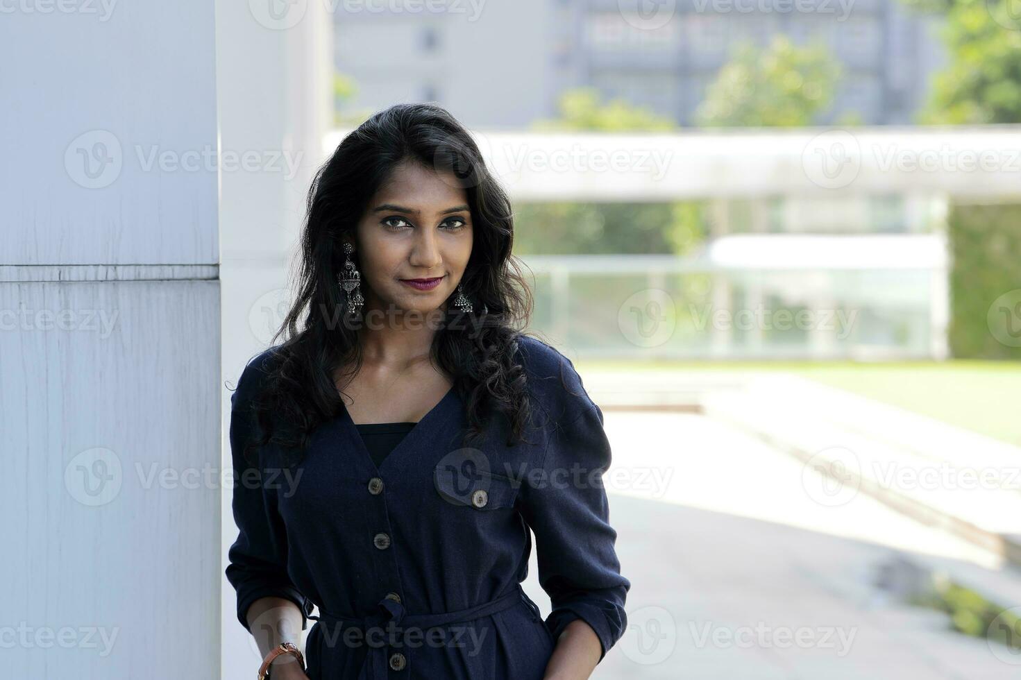 Young attractive beautiful Indian woman in dark blue formal jumpsuit look forward confidently leaning on urban building wall look at camera photo