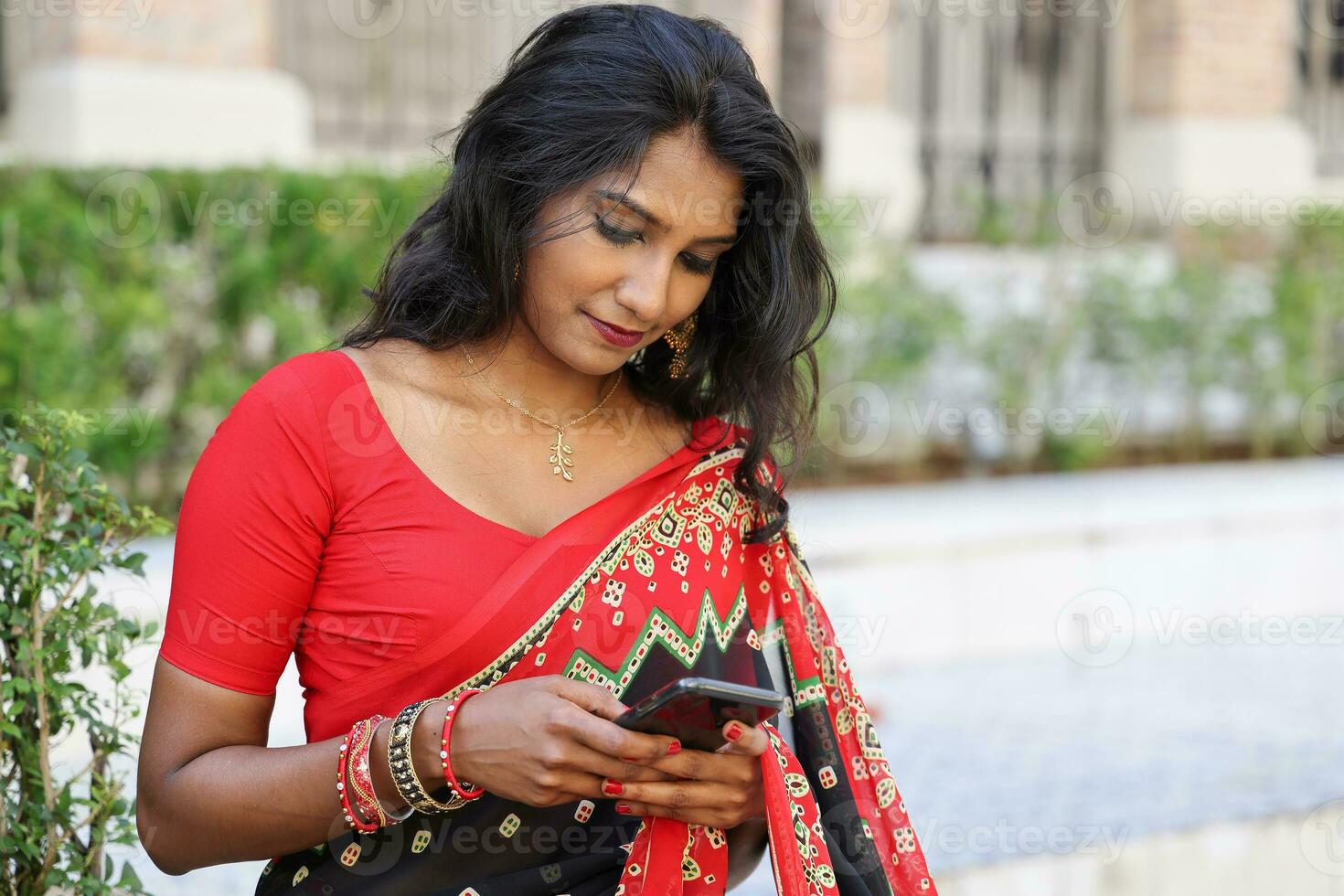 Young attractive beautiful Indian woman in red and black saree texting reading massage smartphone outdoors urban brick wall background photo