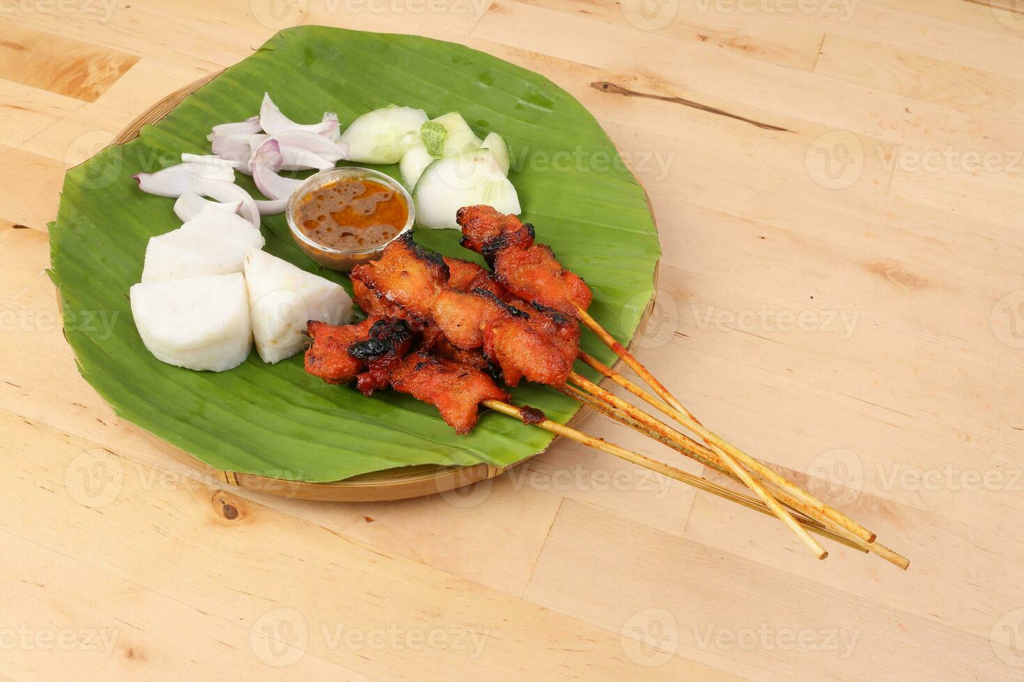 Chicken beef mutton meet Satay set nasi impit sticky rice cake sliced onion cucumber sweet peanut sauce on banana leaf bamboo basket on wooden background photo