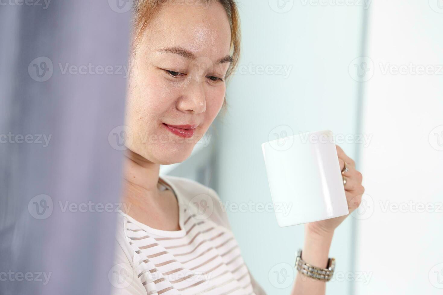 Beautiful attractive middle age southeast Asian woman standing next to daylight window curtain drink coffee from cup looking outside photo