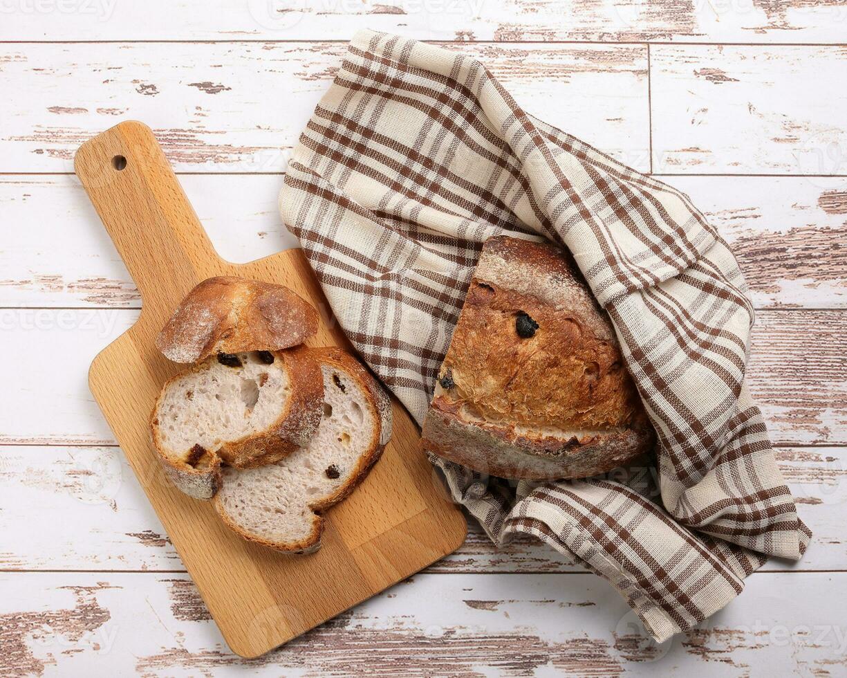 Rustic whole grain artisan bread loaf slice with cranberry raisin dry fruit nuts wrapped in checkers cloth with wooden chopping board over table top flat lay view photo