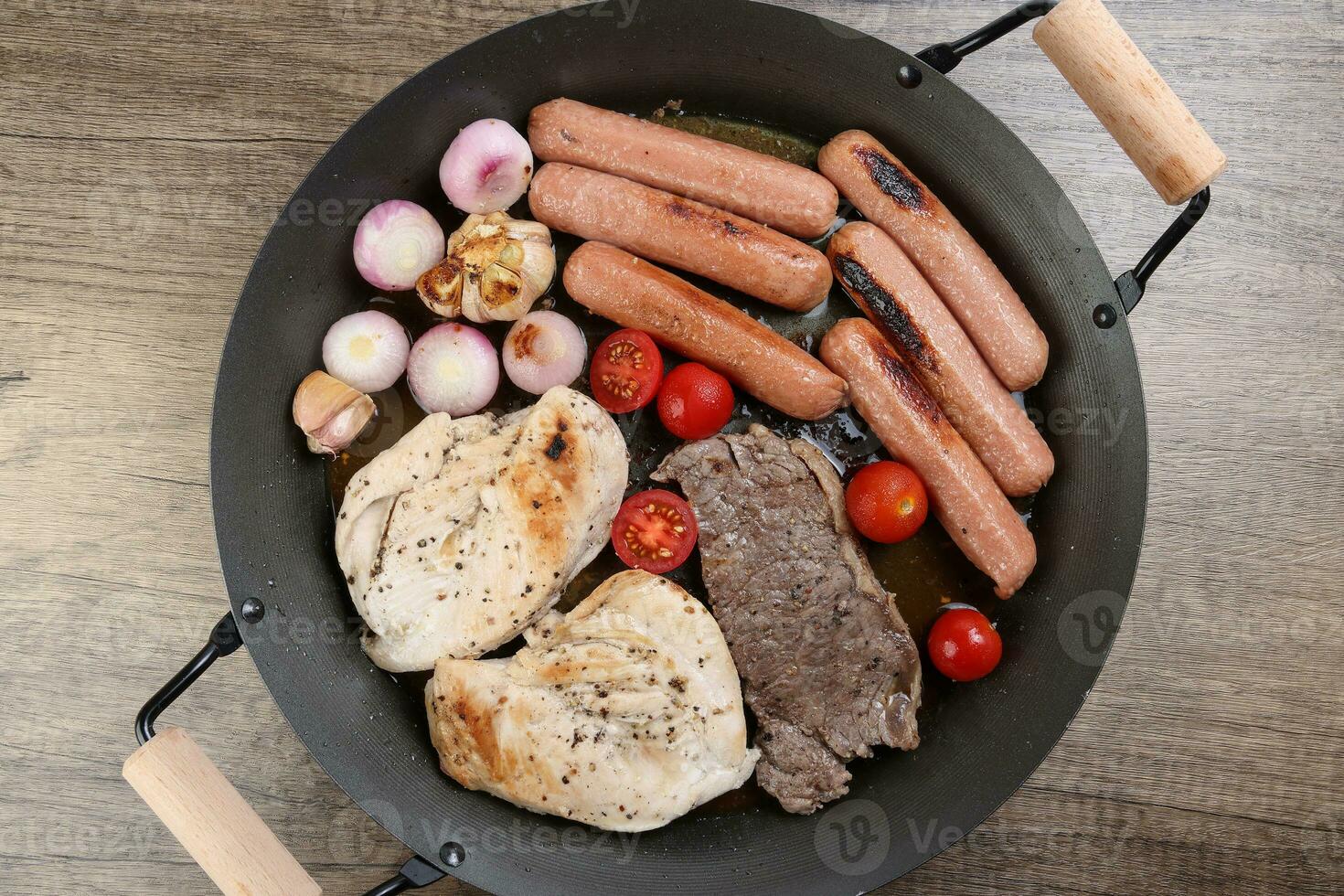 Mix meet pan chicken breast beef steak sausage tomato onion garlic oil fry grill on metal frying pan two wood handle on rustic wooden background photo