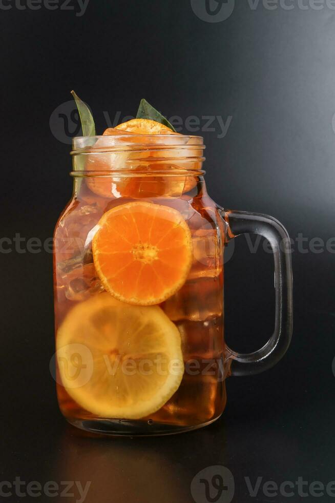 Liquid ice lemon orange tea with slice green leaf cinnamon stick in transparent glass jar mug on black background photo