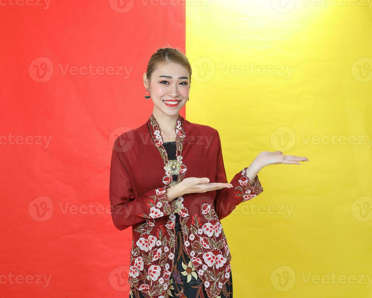 Asian woman traditional maroon kebaya sharong on red yellow paper background posing show display palm of hand photo