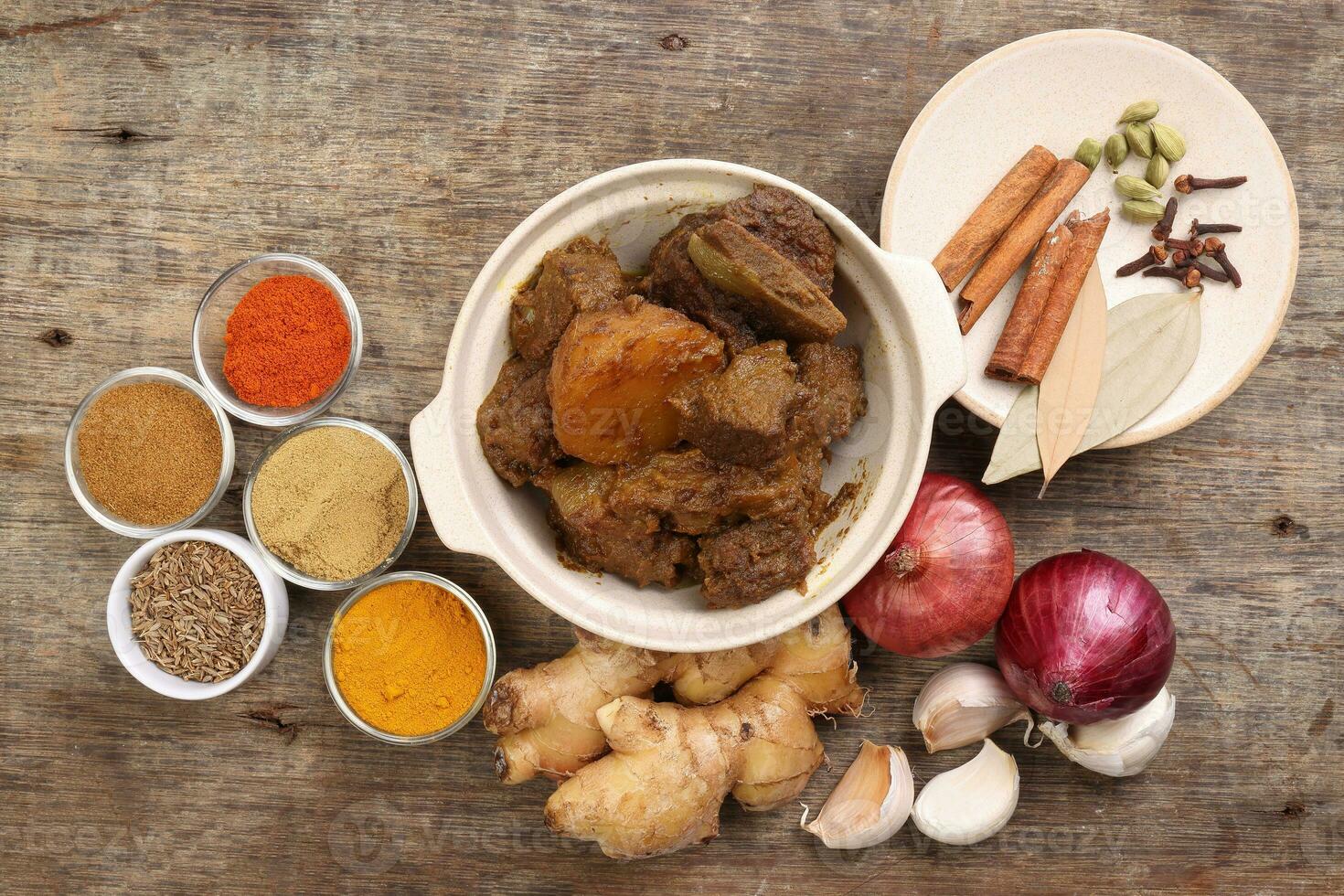 Beef bhuna dry curry with potato in ceramic bowl ingredients chilli cumin coriander turmeric powered salt onion garlic ginger bay leaf cinnamon cardamom clove over rustic wood background photo