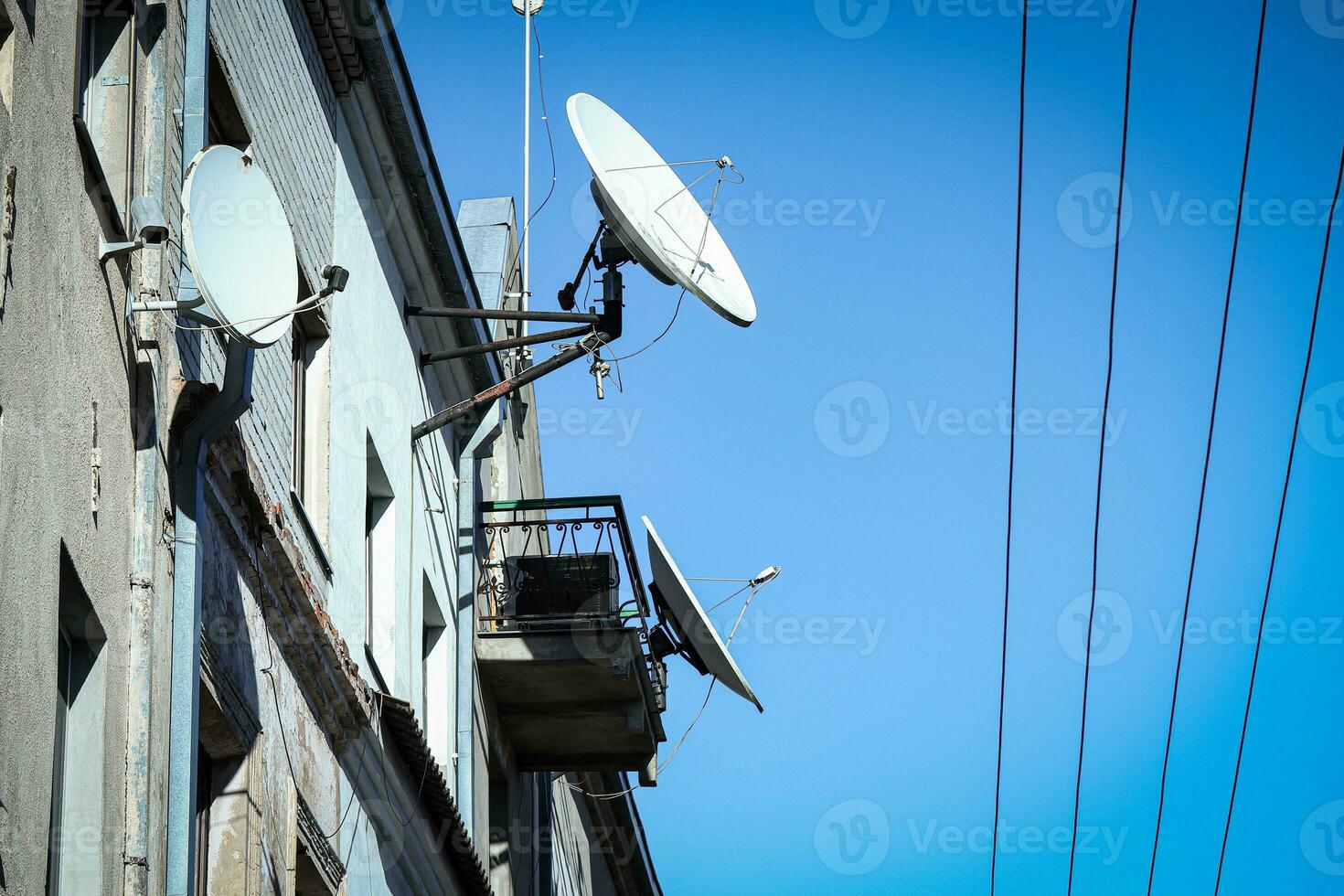 White satellite dish antennas on the roof and wall of ann old building with lines or wires on blue sky background photo
