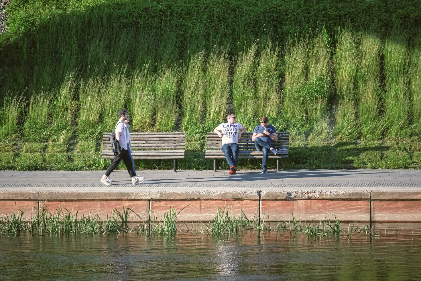 Vilna, Lituania 03 06 2022 dos hombres sentado en un banco y un Pareja caminando a lo largo el río banco en puesta de sol luz de sol foto