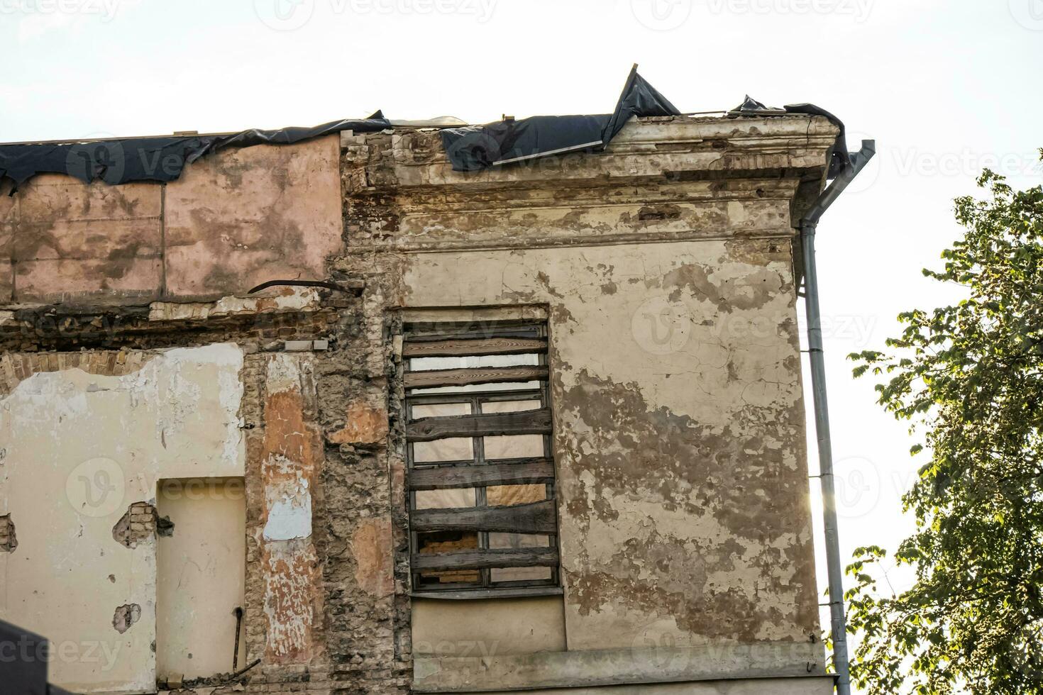 Old window nailed with wooden planks of abandoned aged house corner with peeling plaster on bricks and water pipe photo