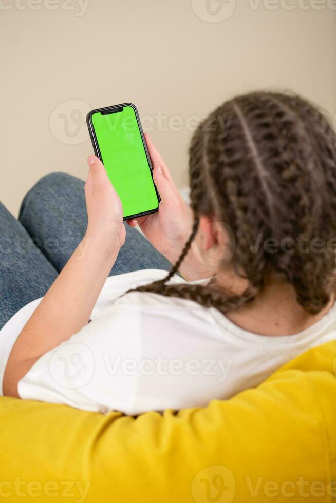 Young girl sitting on a sofa and using a smartphone with green screen photo