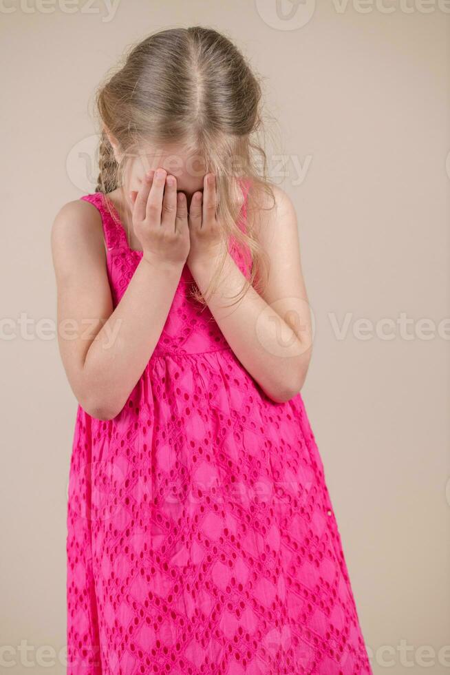 Little girl in a pink dress on a beige background covers her face with her hands photo