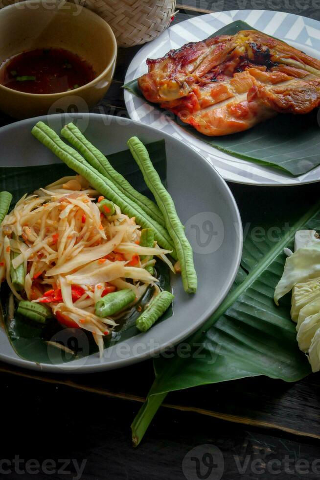 Thai food dish both in Thailand and Asia, Papaya Salad or as we call it Somtum is complemented with grilled chicken and sticky rice with fresh stir-fries. Served on the black wooden table. photo