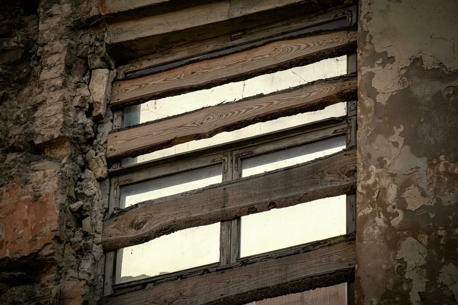 Old window nailed with wooden planks of abandoned aged house with peeling plaster on bricks photo