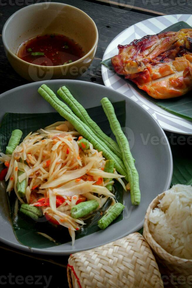 Thai food dish both in Thailand and Asia, Papaya Salad or as we call it Somtum is complemented with grilled chicken and sticky rice with fresh stir-fries. Served on the black wooden table. photo