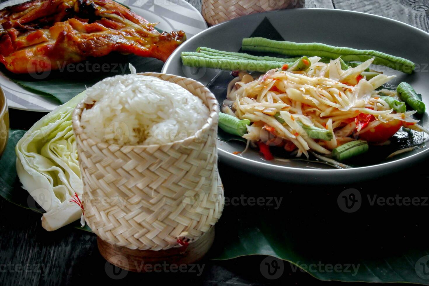 Thai food dish both in Thailand and Asia Papaya Salad or as we call it Somtum is complemented with grilled chicken and sticky rice with fresh stir-fries. Served on the black wooden table. photo