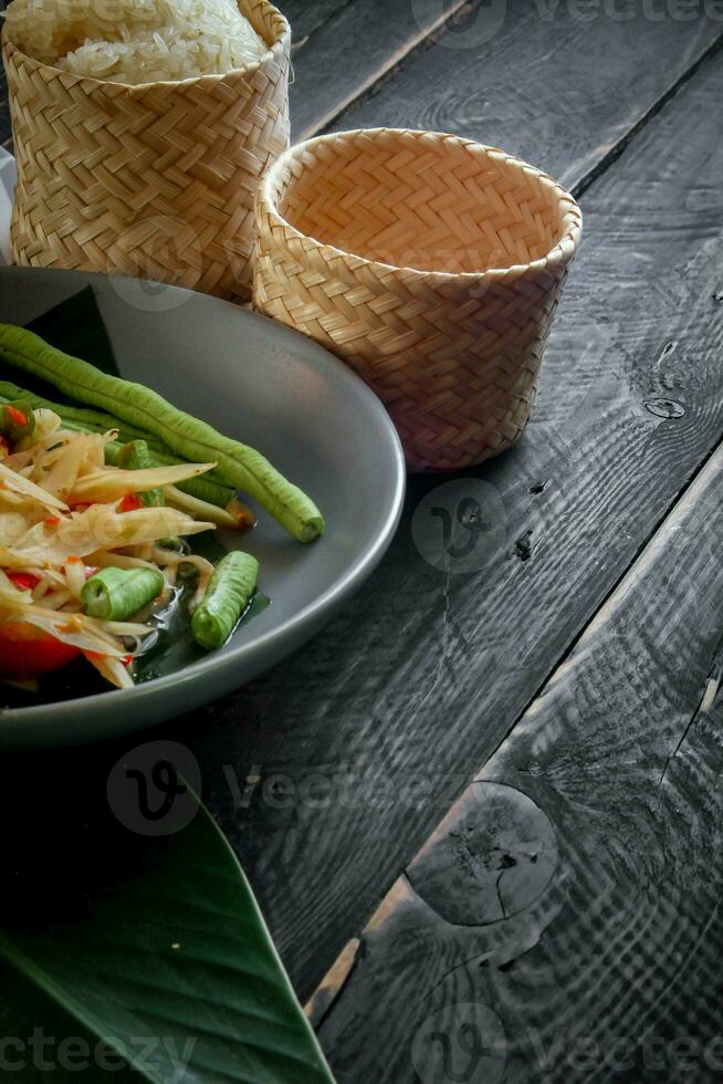 Thai food dish both in Thailand and Asia, Papaya Salad or as we call it Somtum is complemented with grilled chicken and sticky rice with fresh stir-fries. Served on the black wooden table. photo