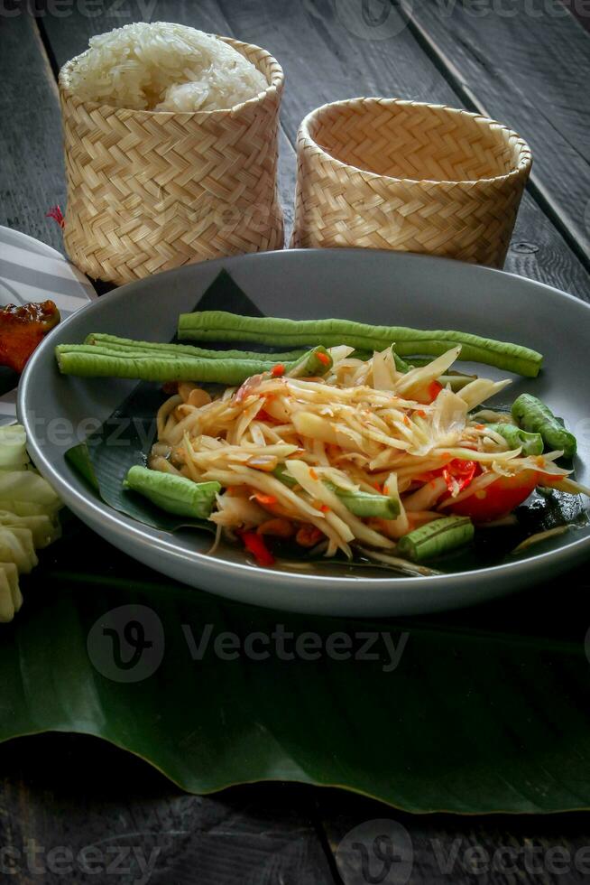 Thai food dish both in Thailand and Asia, Papaya Salad or as we call it Somtum is complemented with grilled chicken and sticky rice with fresh stir-fries. Served on the black wooden table. photo