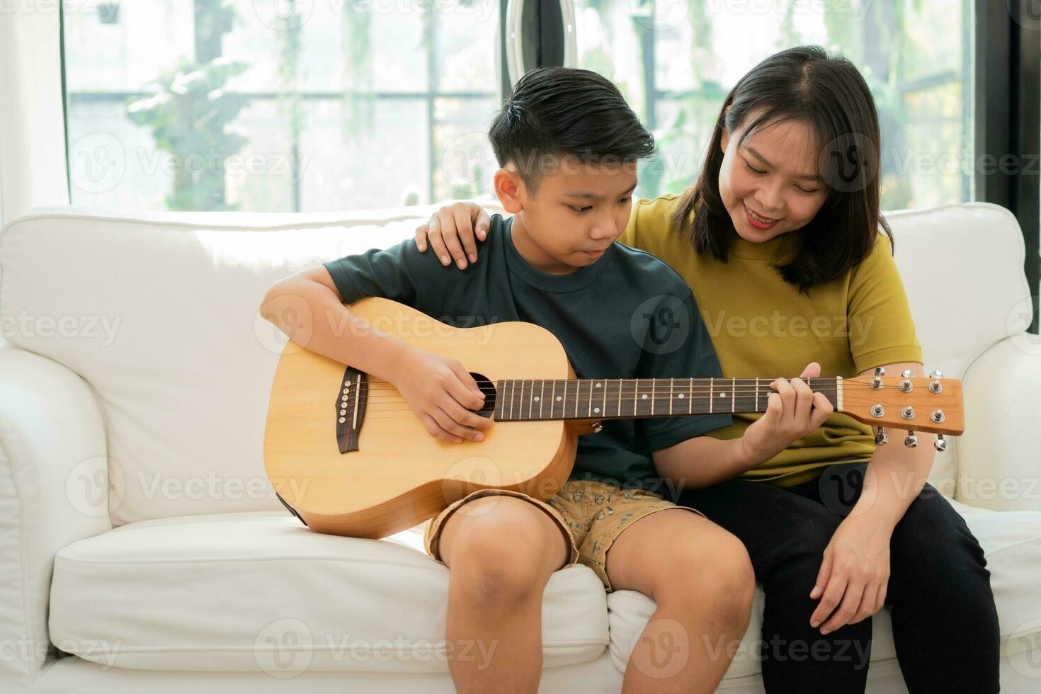 la madre asiática abraza al hijo, el niño asiático tocando la guitarra y la madre se abrazan en el sofá y se sienten apreciados y alentados. concepto de una familia feliz, aprendizaje y estilo de vida divertido, amar los lazos familiares foto