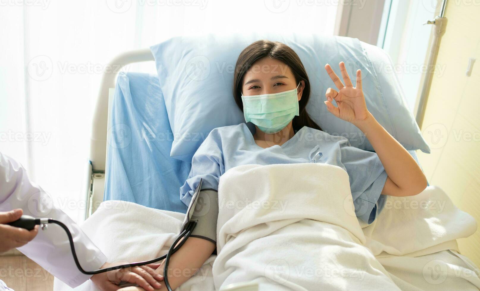 Happy Asian female patient wears a mask, lies on the bed, and Shows an ok symbol. When the doctor examines the symptoms using a pressure gauge. Concept of believe in treatment And insurance coverage photo