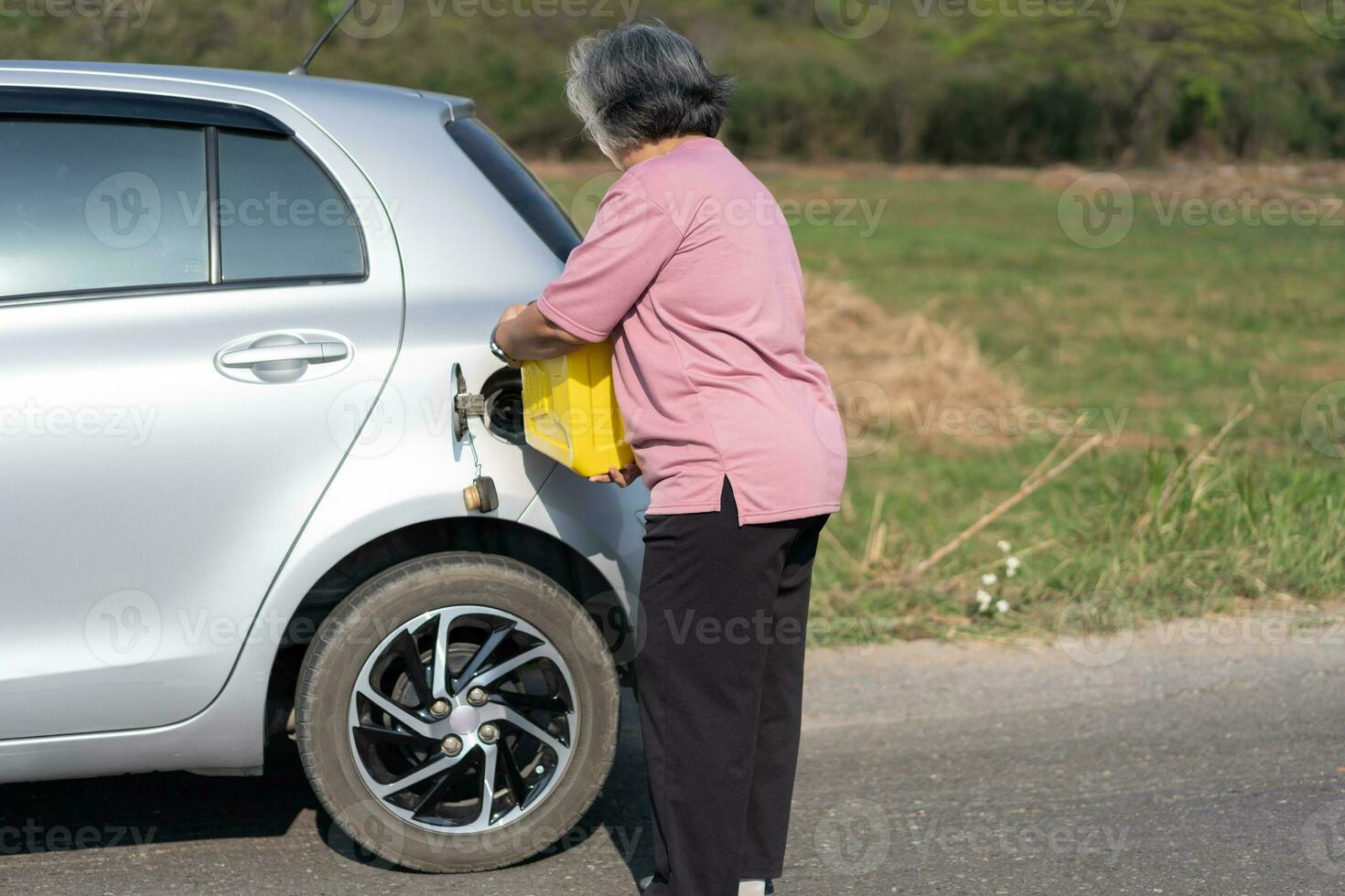 The car ran out of gas and stalled beside the road in suburbs and an elderly Asian woman used a gallon of spare gas to fuel the car. A woman prepares a gallon of spare gas to fuel before traveling. photo