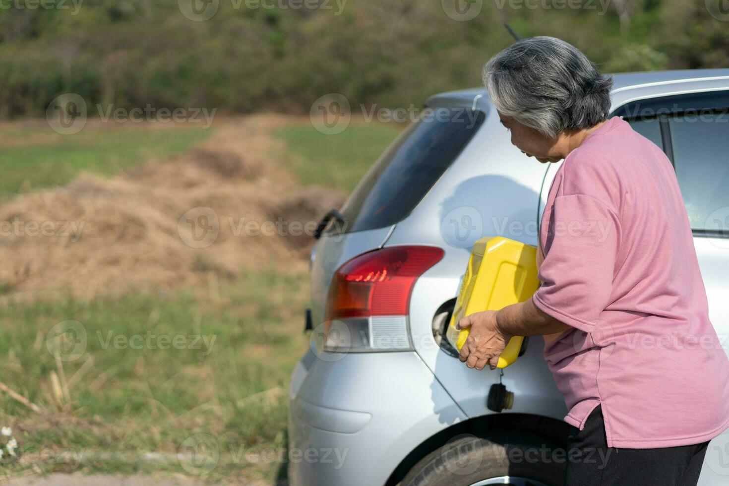 The car ran out of gas and stalled beside the road in suburbs and an elderly Asian woman used a gallon of spare gas to fuel the car. A woman prepares a gallon of spare gas to fuel before traveling. photo