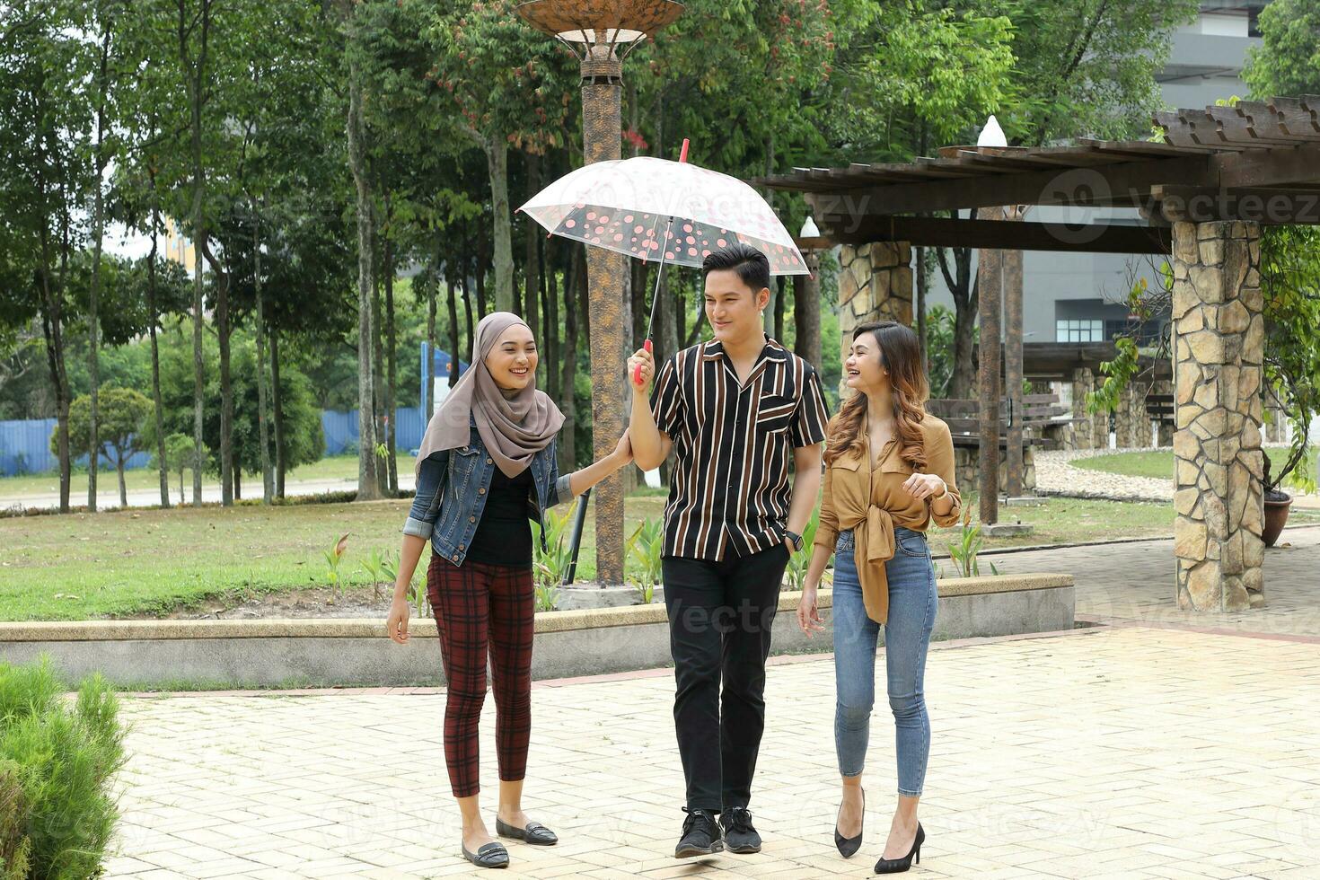 Young Asian Malay man woman outdoor green park walk talk discuss mingle under umbrella photo
