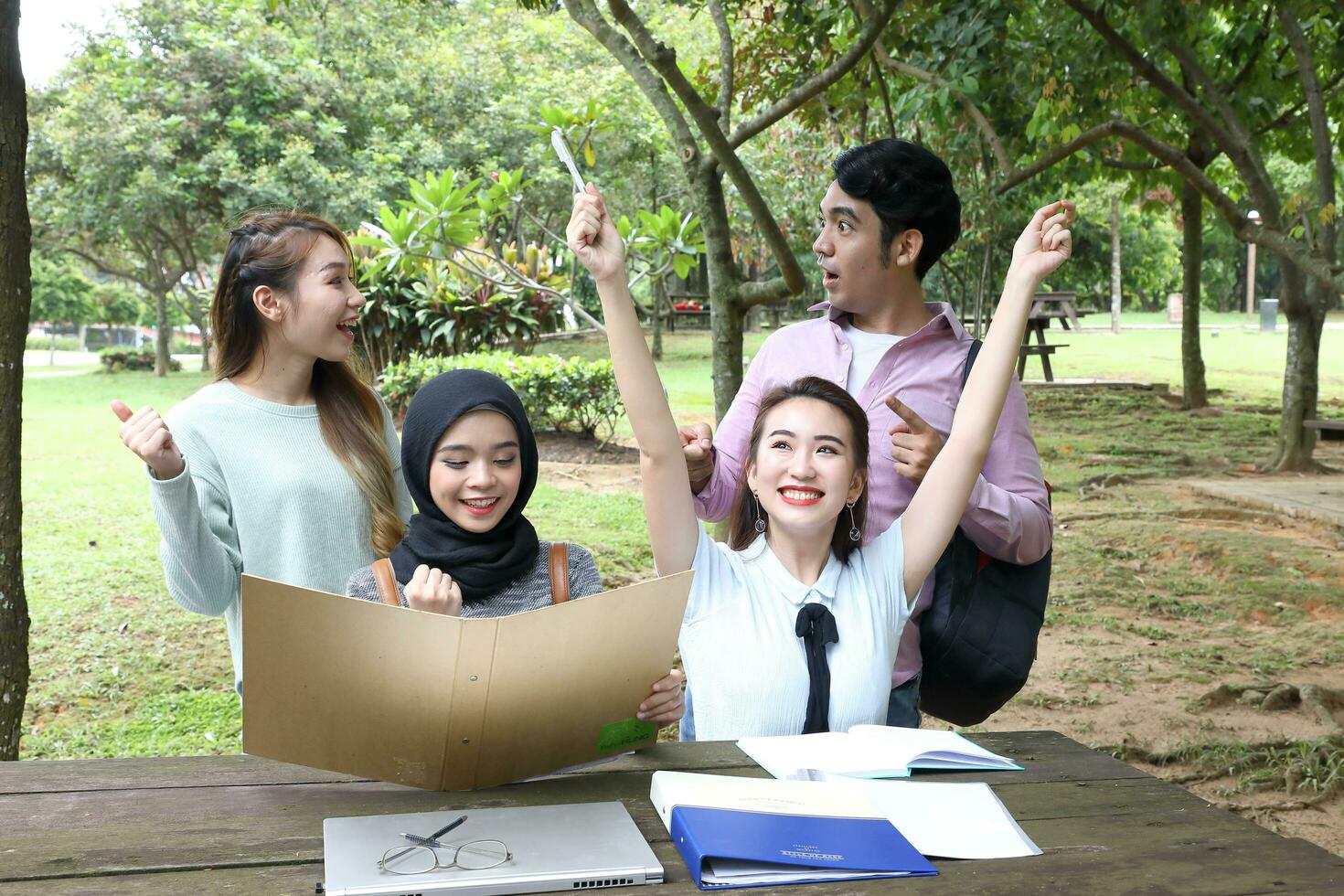 Young asian malay chinese man woman outdoor on park bench table study talk discuss point laptop file book happy hand in the air photo