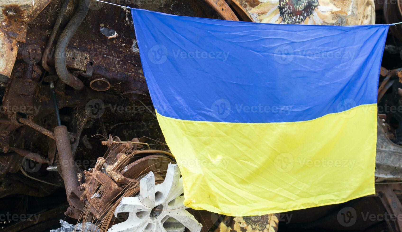 The national flag of Ukraine against the background of ruins and rusty metal of destroyed cars. War in Ukraine. A blue-yellow flag waving in the wind under the sun in a bomb-ravaged city. photo