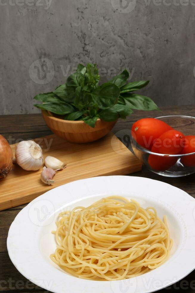 Cooked spaghetti on white plate with ingredients roma tomato glass bowl basil onion garlic wooden board recipe on table marble wall photo