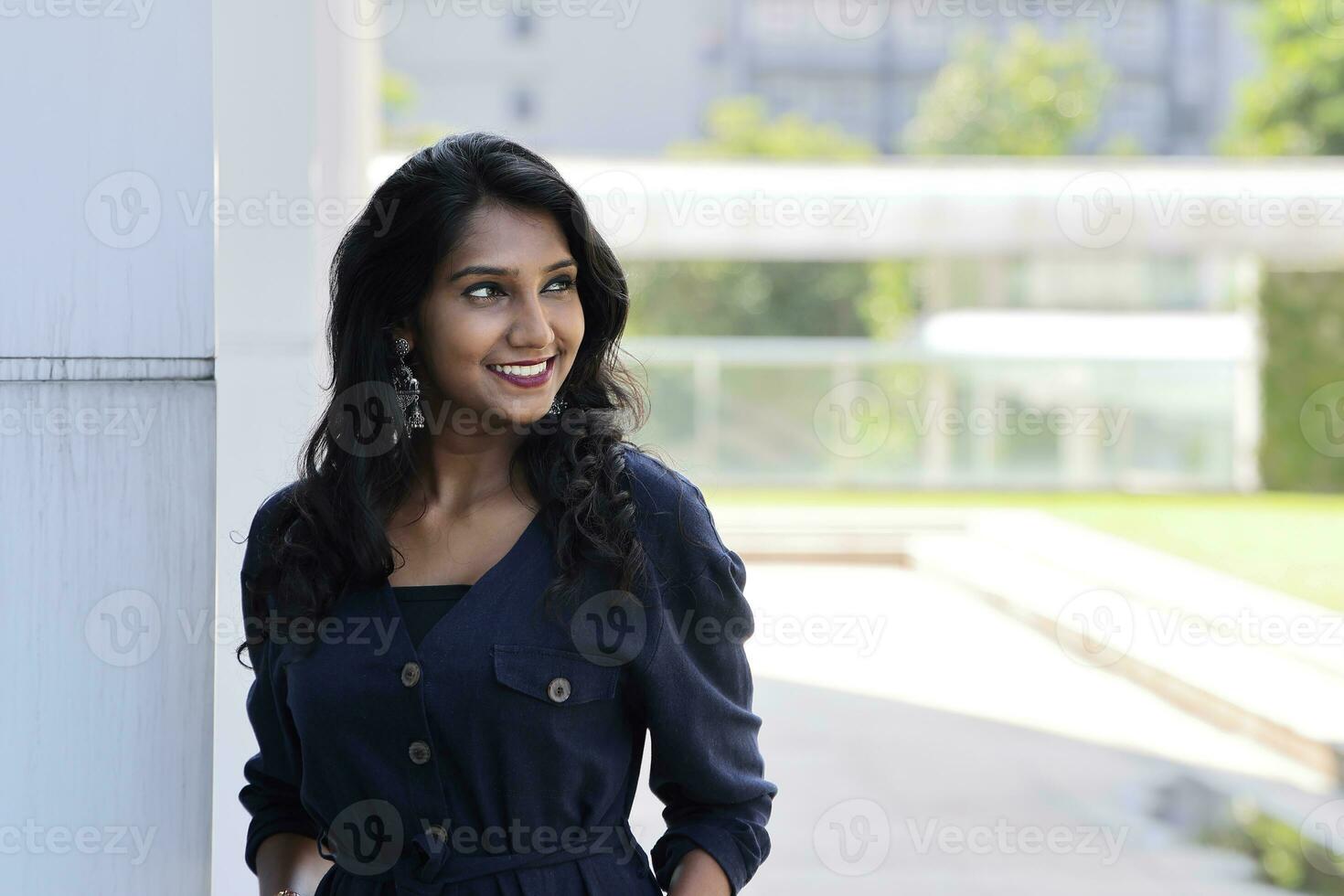 Young attractive beautiful Indian woman in dark blue formal jumpsuit look forward confidently leaning on urban building wall look away smiling photo