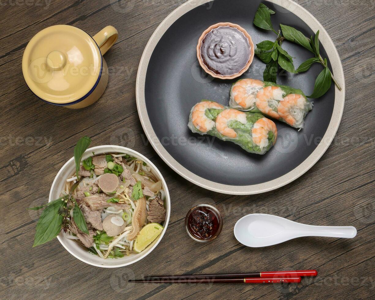 Vietnamese prawn shrimp see through rice paper spring roll poh beef noodles soup beef ball brisket tripe stomach parts basil lime rustic wooden table chopsticks spoon sauce dark plate tin mug photo