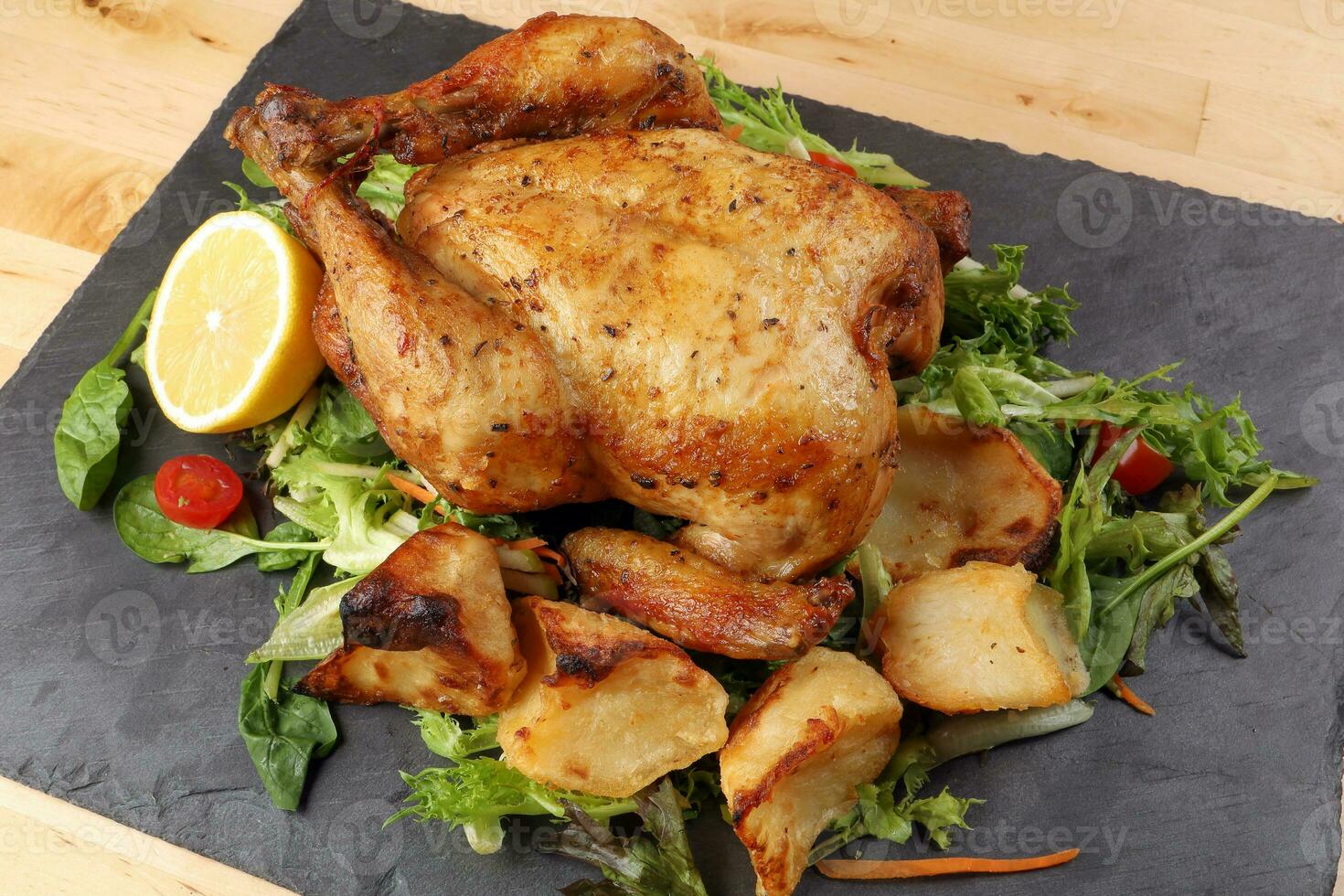 Whole roasted grilled chicken poultry bird with baked potato vegetable salad tomato lemon on black slate stone cutting board wooden table background photo