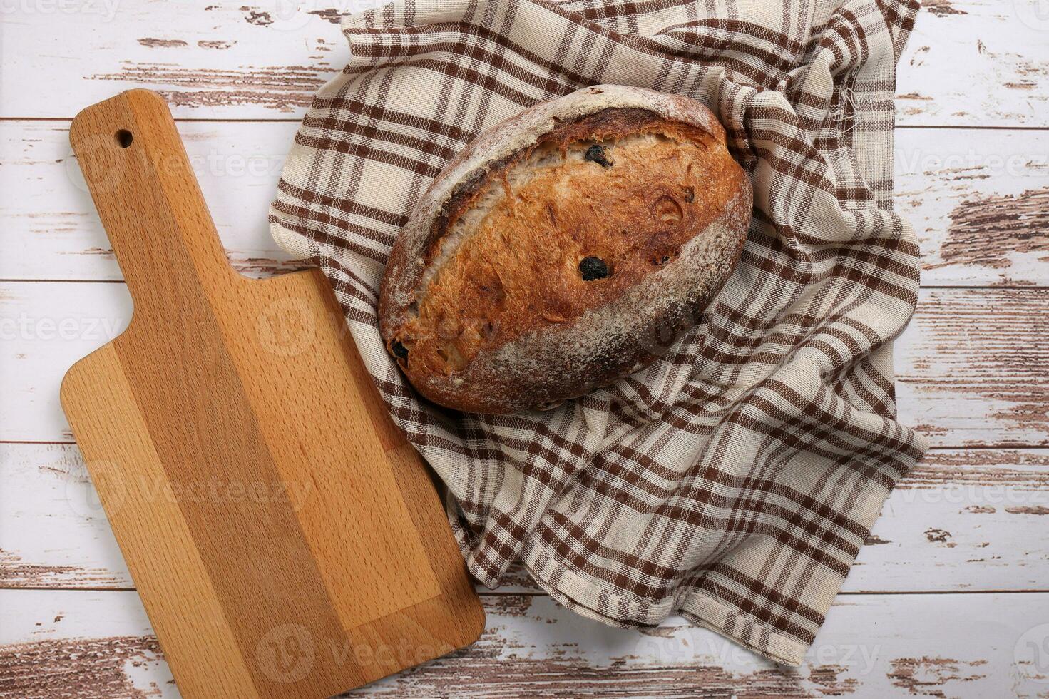 Rustic whole grain artisan bread loaf with cranberry raisin dry fruit nuts wrapped in checkers cloth with wooden chopping board over table top flat lay view photo