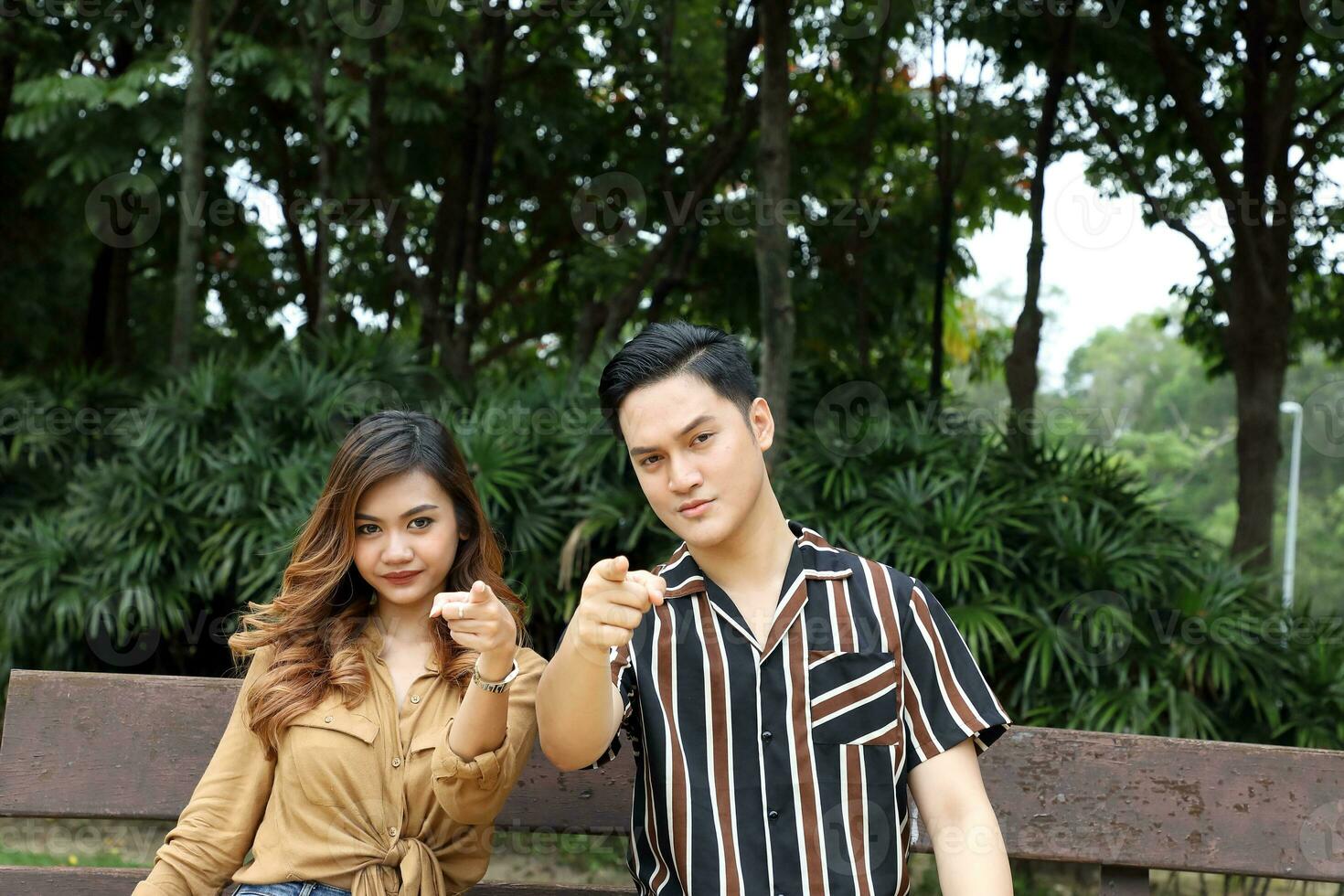 oung Asian Malay man woman couple outdoor green park sit on bench point finger front at camera photo