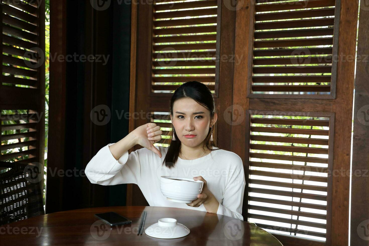 Young asian woman sit at rustic cafe holding soup noodle bowl hand finger thumbs down bad sign look front at camera photo