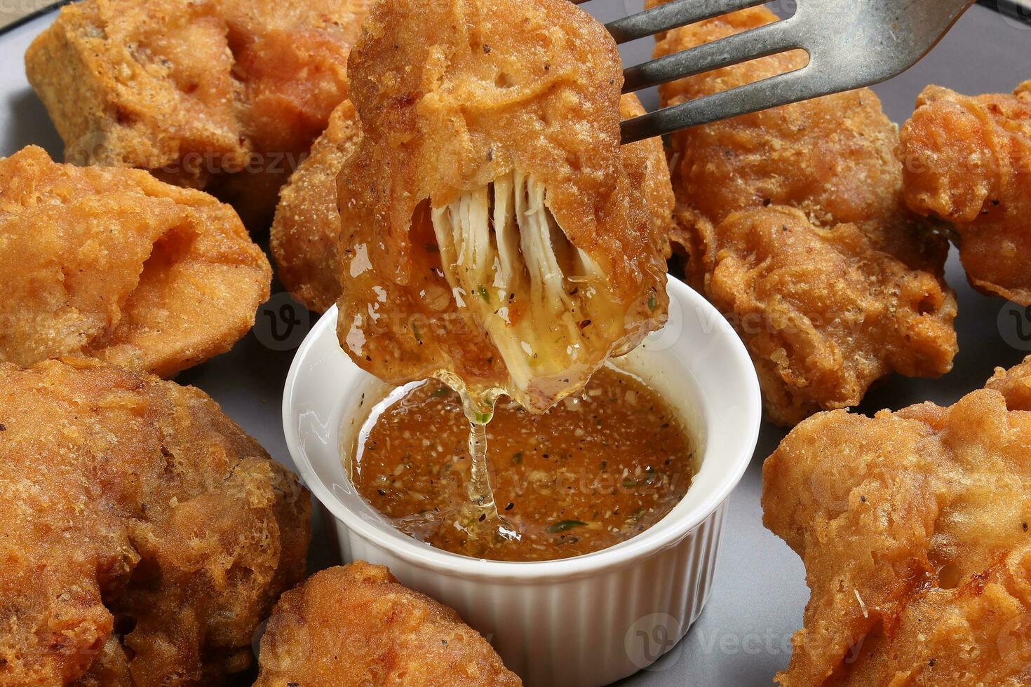 Light battered deep fried chicken variety mix pieces pile flaky meat on grey plate on fork dripping honey garlic clear sauce in center over wooden background photo