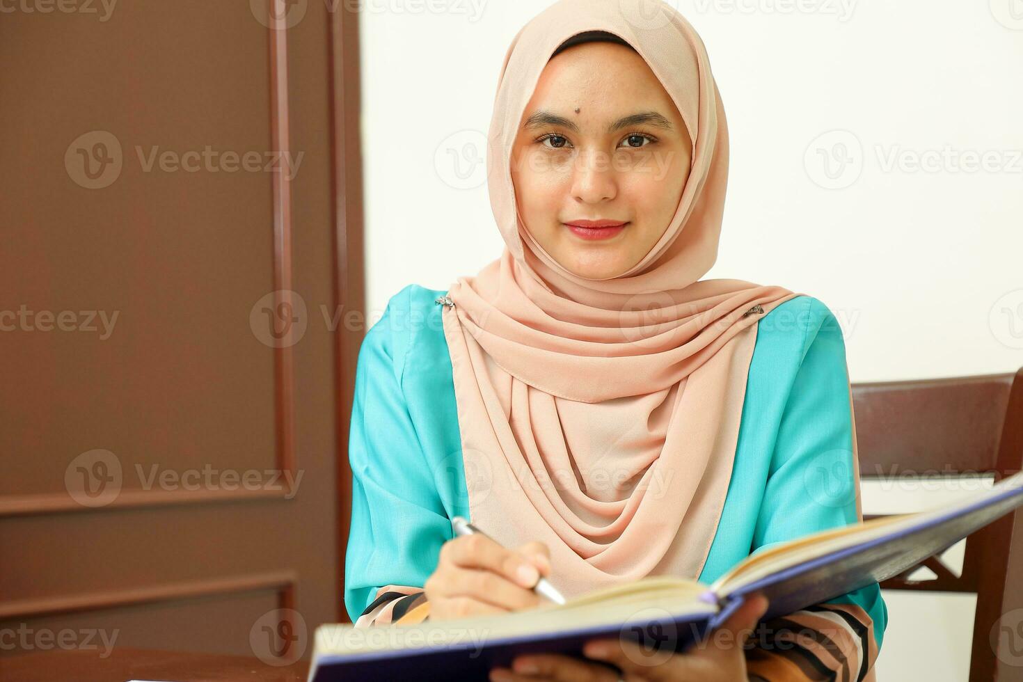 young Asian Malay Muslim woman wearing headscarf at home office student sitting at table read write book document note study smile happy look at camera photo