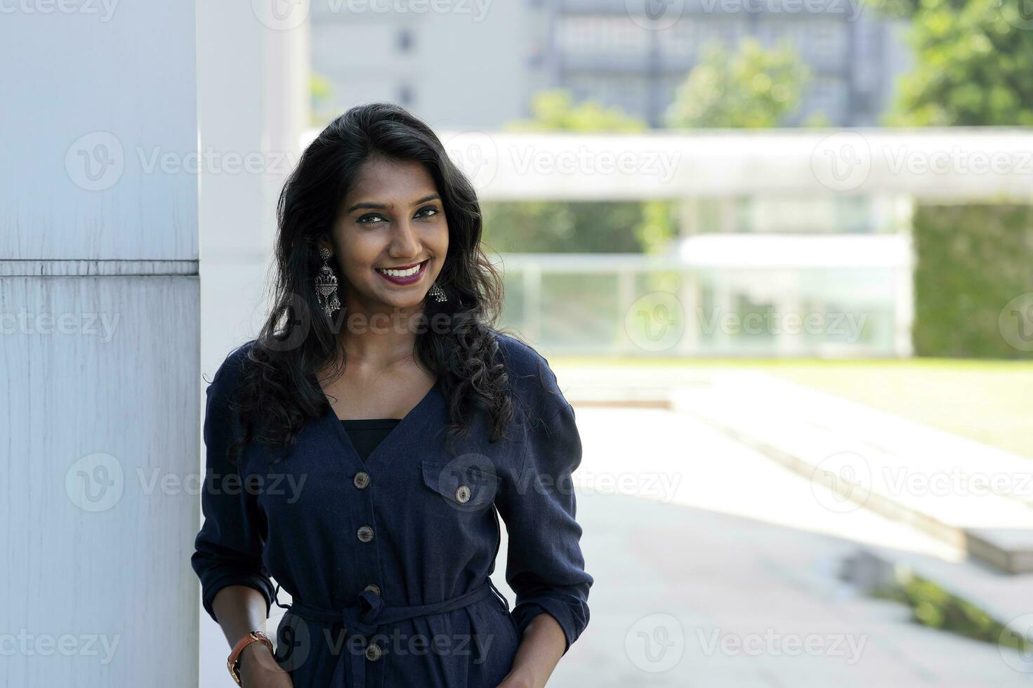 Young attractive beautiful Indian woman in dark blue formal jumpsuit look forward confidently leaning on urban building wall look at camera smiling photo