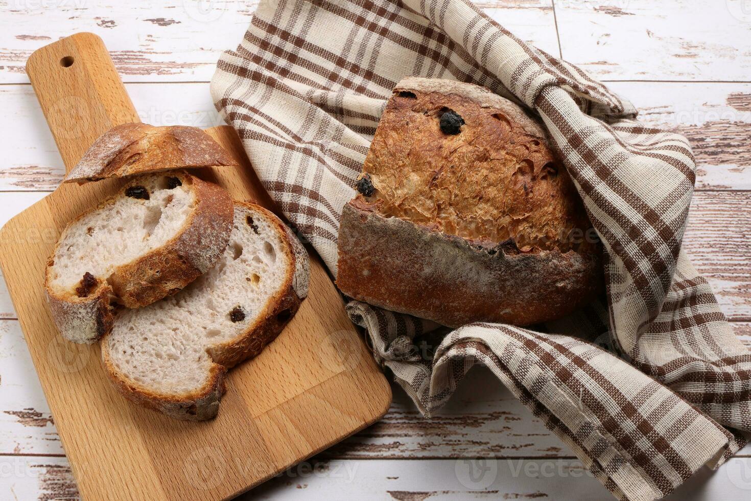 Rustic whole grain artisan bread loaf slice with cranberry raisin dry fruit nuts wrapped in checkers cloth with wooden chopping board over table top flat lay view photo