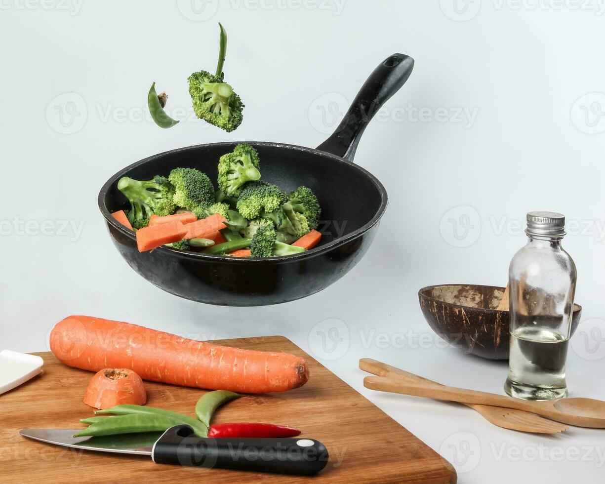 chopped sliced diced carrot broccoli white button mushroom red chili flying dropping gravity defying on floating elevated metal frying pan cooking board bowl oil knife on white background photo